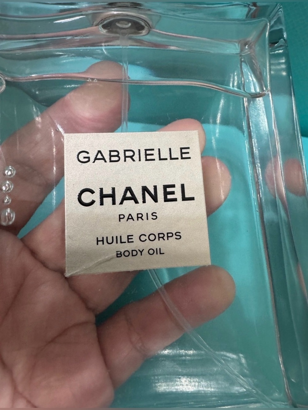 CHANEL Gabrielle Body Oil in Clear Bottle with Teal Background
